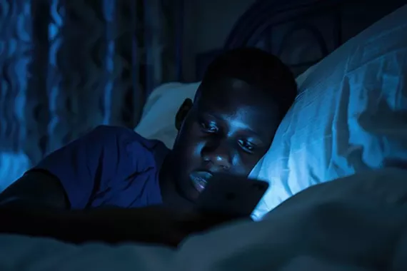 A teen boy in bed in a dark bedroom. He is on his side propped up on a pillow. He is looking at his cellphone, which shines on his face
