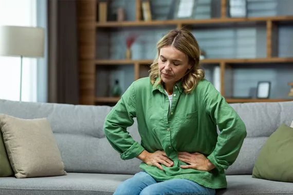 A young woman is sitting on a couch and has both hands on her stomach. She has her eyes closed and looks very uncomfortable