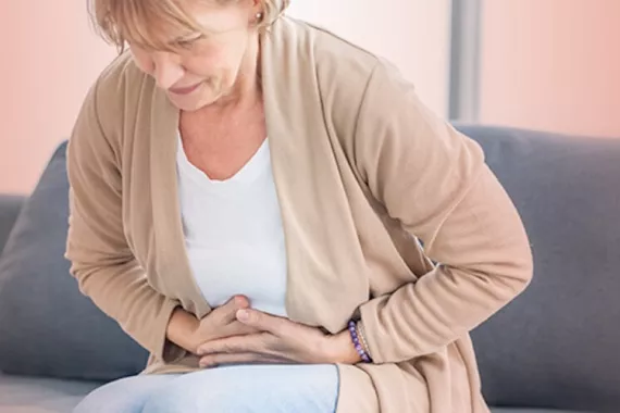 A middle-aged woman is sitting on a couch. She is leaning forward and has both hands on her stomach as if she is having intense stomach pain