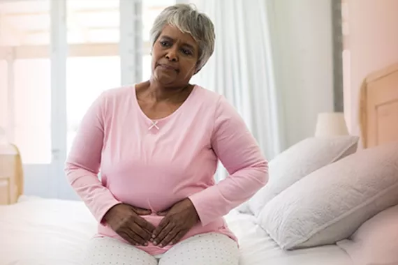 An older woman sits on her bed while grabbing her abdomen with a pained expression on her face