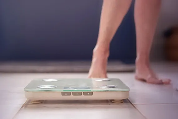 A digital scale sits on the floor in the foreground, in the background a pair of legs indicates a person is about to step onto the scale