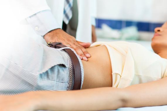 A doctor examines a young female patient lying on an exam table in a doctor’s office. The doctor is touching the patient’s abdomen