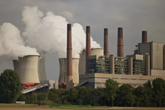 Industrial site with seven smokestacks. Four are tall and thin and three are large, wide and cylindrical. White smoke gushes into the air
